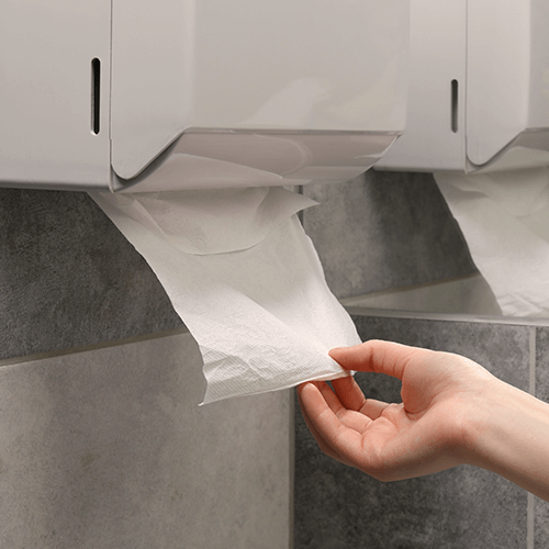 Eine Hand zieht Papiertücher aus einem Papiertuchspender, der an einer Wand hängt